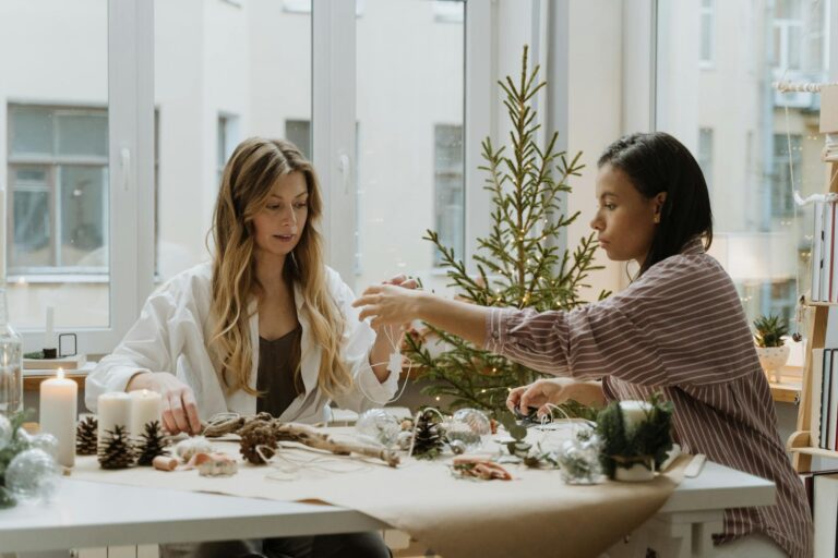 Two women engaging in a creative holiday crafting process indoors with candles and festive decor.
